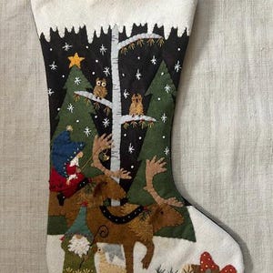 Puede incluir: Un calcetín navideño con una escena de bosque invernal. El diseño presenta un fondo negro con árboles verdes, búhos y una estrella. También se representan un gnomo, un reno y una figura con un sombrero azul. La parte superior del calcetín es blanca.