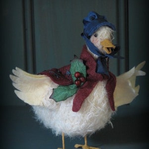 May include: A white, fluffy goose wearing a red and burgundy cape, a blue hat, and a green holly sprig with red berries. The goose has a yellow beak and feet.