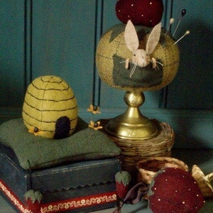 May include: A set of three needlepoint pincushions. One pincushion is shaped like a beehive and sits on a green box with a red and white trim. Another pincushion is shaped like a strawberry and sits on a gold stand. The third pincushion is shaped like a strawberry and is sitting on a table.
