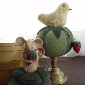 May include: A green felt pincushion shaped like a tomato with a white fluffy bird perched on top. A brown felt mouse with a red felt strawberry is sitting on a green felt pincushion.
