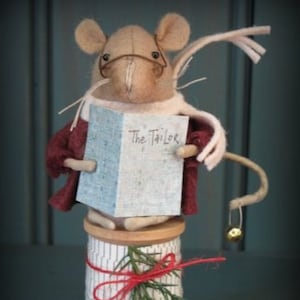 Puede incluir: Un ratón de fieltro con gafas y una bufanda rosa sostiene un libro que dice "The Tailor". El ratón está sentado en un carrete de madera con una cinta roja y ramitas de pino verde.