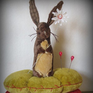 May include: A brown plush rabbit with long ears holds a white flower with a red center. The rabbit is sitting on a green pincushion with red stitching. There are two red pins sticking out of the pincushion.