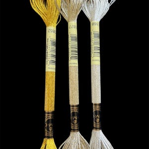 May include: Three spools of metallic embroidery floss in gold, silver, and off-white. Each spool has a black label with white text and a barcode. The floss is tightly wound at the base and flares out at the top. The background is black.