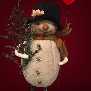 Könnte beinhalten: Ein skurriler Schneemann mit schwarzem Zylinder, braunem Schal und Karottennase. Er hält einen Zweig Grünzeug und ein rotes Herz an einem Stock. Der Schneemann hat goldfarbene Knöpfe und eine Blume auf dem Hut. Der Hintergrund ist rot.