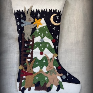 May include: A felt Christmas stocking with a dark blue background and a white cuff. The stocking features a green Christmas tree with white snowflakes, a crescent moon, and three brown rabbits decorating the tree. One rabbit is climbing a ladder, while the other two are holding ornaments.