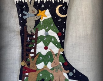 May include: A felt Christmas stocking with a dark blue background and a white cuff. The stocking features a green Christmas tree with white snowflakes, a crescent moon, and three brown rabbits decorating the tree. One rabbit is climbing a ladder, while the other two are holding ornaments.