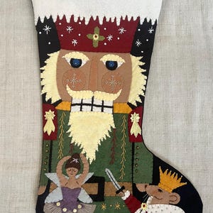 Puede incluir: Una media de Navidad de fieltro con un diseño de Cascanueces. La media presenta un sombrero rojo, una chaqueta verde, una barba blanca y una espada. Una bailarina y un rey ratón también están representados en la media.