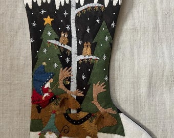 Puede incluir: Un calcetín navideño con una escena de bosque invernal. El diseño presenta un fondo negro con árboles verdes, búhos y un alce. También hay un gnomo y una persona con un abrigo rojo. La parte superior del calcetín es blanca.