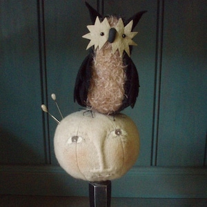 May include: A whimsical owl with black wings and a brown, fluffy body sits atop a white, round pincushion with a face. The pincushion is on a black pedestal.