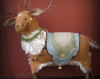 May include: A brown felt reindeer with a twig antler, wearing a blue, green, and white felt saddle with a gold chain and bell. The reindeer is standing on a dark surface.