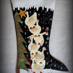 Peut inclure: Une chaussette de Noël en feutre noir avec un revers blanc. Un sapin de Noël en feutre vert avec des guirlandes lumineuses rouges est sur le côté gauche de la chaussette. Une pile de cinq oies en feutre blanc avec des accents en feutre brun sont sur le côté droit de la chaussette. La chaussette porte les mots "Joy to the World!" brodés dessus.