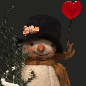 May include: A handcrafted snowman figure with a black top hat adorned with a peach-colored flower. The snowman has a carrot nose, a brown scarf, and a gold brooch. It holds a red heart-shaped balloon and a sprig of greenery.