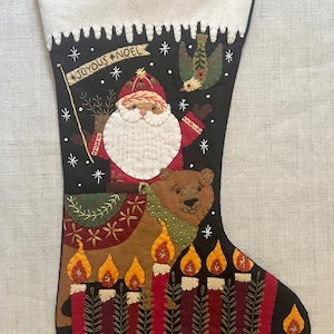 May include: A black Christmas stocking with a white cuff, featuring Santa Claus riding a bear. The stocking has a banner that says "JOYOUS NOEL" and is decorated with candles, snowflakes, and a bird.