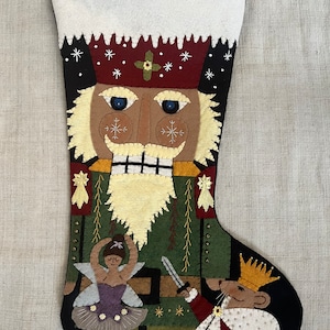 May include: A felt Christmas stocking with a Nutcracker design. The stocking features a green and brown Nutcracker with a white beard, a red hat, and a blue face. A ballerina doll and a mouse king are also depicted on the stocking.