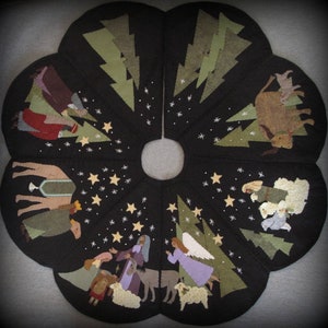 May include: A felt Christmas tree skirt with a black background and a depiction of the nativity scene. The skirt features a camel, a donkey, a shepherd, an angel, and a star. The scene is surrounded by green trees and white stars.