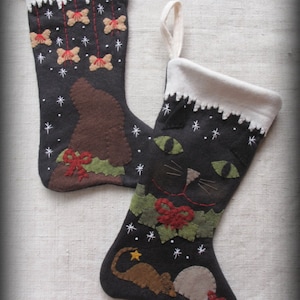 Puede incluir: Dos calcetines de Navidad de fieltro, uno con un gato negro y el otro con un perro marrón. Ambos calcetines tienen un puño blanco y están decorados con copos de nieve y otros detalles festivos.