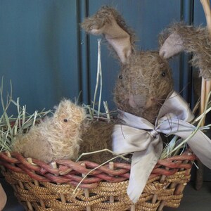 May include: A brown and white bunny rabbit and a small brown and white chick are sitting in a woven wicker basket filled with straw. The bunny has a silver ribbon tied around its neck.
