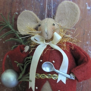 May include: A felt mouse ornament wearing a red coat, a white ribbon, and a gold trim. The mouse is holding a silver spoon and has a green sprig of greenery in its other hand.
