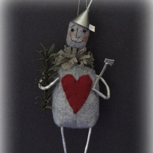 Puede incluir: Un adorno de fieltro gris del Hombre de Hojalata de El Mago de Oz, sosteniendo un pequeño hacha y un corazón rojo. Lleva un sombrero plateado y tiene una cara amigable.