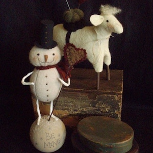 May include: A white felt snowman with a black top hat and a red scarf stands on a white ball that says "Be Mine". A white felt sheep with a red heart on its side stands behind the snowman.