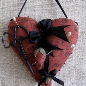 Könnte beinhalten: Ein rotes herzförmiges Nadelkissen mit schwarzem Band und einer Schere. Das Nadelkissen ist mit einem Blumenmuster verziert und hat mehrere Nadeln, die herausragen.
