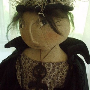 May include: A handmade rag doll wearing a black hat with a brown and white trim, a black jacket, and a brown and white patterned dress. The doll has a black necklace with a large pendant.