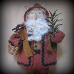 Könnte beinhalten: Ein handgefertigter Weihnachtsbaumschmuck aus Filz, der eine Weihnachtsmannfigur darstellt. Er trägt einen roten Mantel mit schwarzem Besatz, einen braunen Schal und einen weißen Bart. Er trägt einen Korb und ein Bündel Reisig.