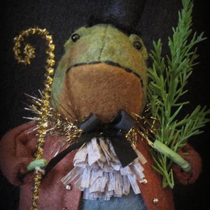 May include: A green frog wearing a top hat, a red jacket, a white ruffle, and a black bow tie. The frog is holding a gold-trimmed stick and a green sprig of foliage.