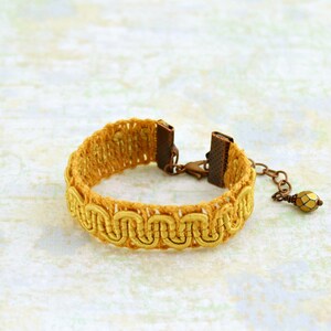 Puede incluir: Un brazalete trenzado amarillo con un cierre de metal y un peque&ntilde;o abalorio de cuentas doradas.