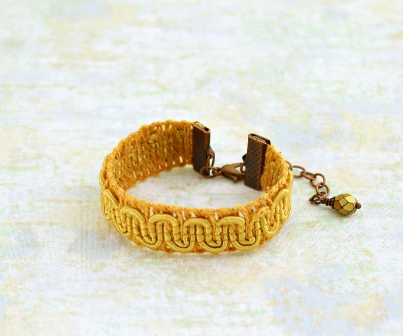 Puede incluir: Un brazalete trenzado amarillo con un cierre de metal y un peque&ntilde;o abalorio de cuentas doradas.