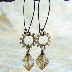 Puede incluir: Un par de pendientes de bronce con un dise&ntilde;o de engranaje y cuentas de cristal transparentes. Los pendientes cuelgan de un alambre.