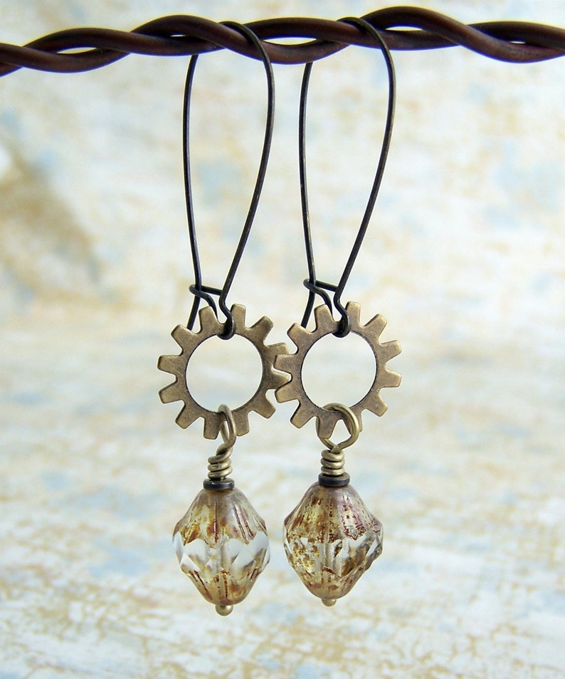 Puede incluir: Un par de pendientes de bronce con un dise&ntilde;o de engranaje y cuentas de cristal transparentes. Los pendientes cuelgan de un alambre.