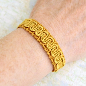 Puede incluir: Un brazalete trenzado dorado con un patr&oacute;n decorativo. El brazalete est&aacute; hecho de tejido tejido y tiene un dise&ntilde;o simple.