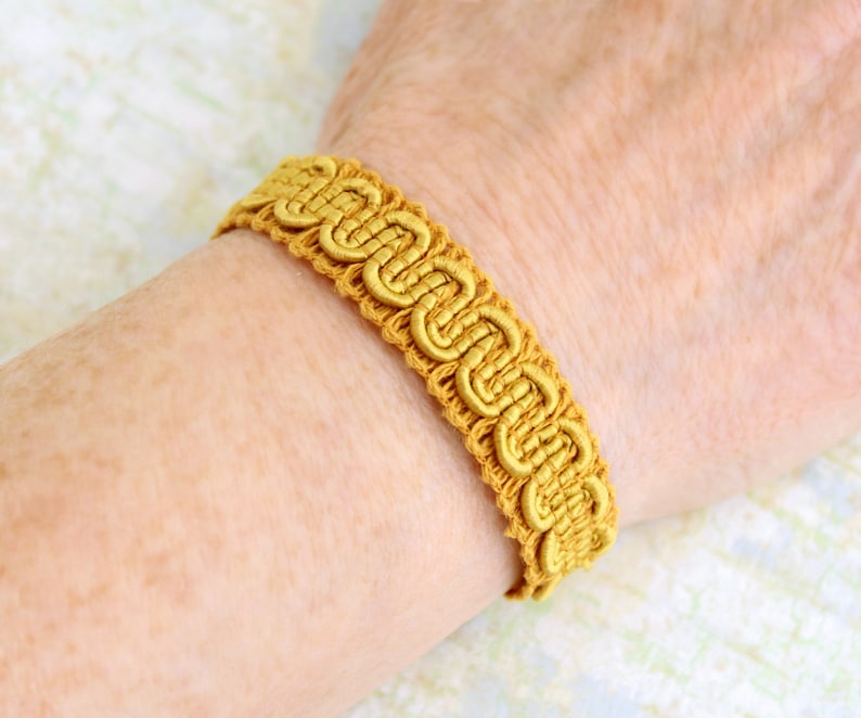 Puede incluir: Un brazalete trenzado dorado con un patr&oacute;n decorativo. El brazalete est&aacute; hecho de tejido tejido y tiene un dise&ntilde;o simple.