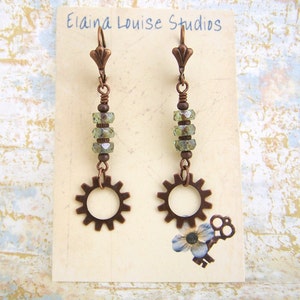 May include: A pair of copper-toned earrings with a gear charm and green crystal beads. The earrings are on a white card with the text "Elaina Louise Studios".