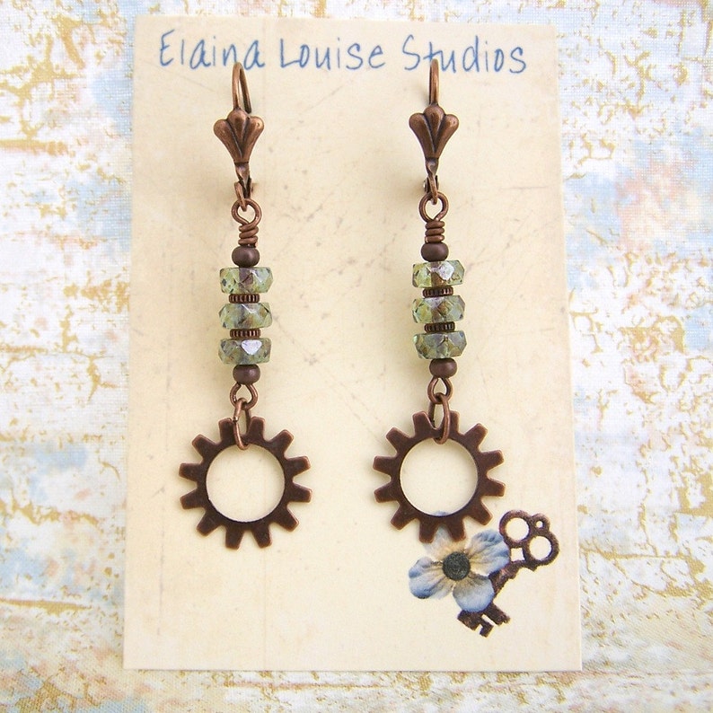 May include: A pair of copper-toned earrings with a gear charm and green crystal beads. The earrings are on a white card with the text "Elaina Louise Studios".