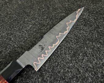 May include: A kitchen knife with a dark, textured steel blade featuring a prominent, wavy copper-colored inlay running along the length of the edge. The handle is constructed from a dark, polished material with a reddish-brown wood grain section.