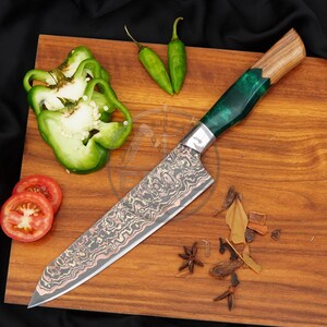 May include: A chef knife with a patterned Damascus steel blade and a handle made of wood and translucent green resin. The knife is positioned next to sliced green bell peppers, two small green chilli peppers, and scattered spices.