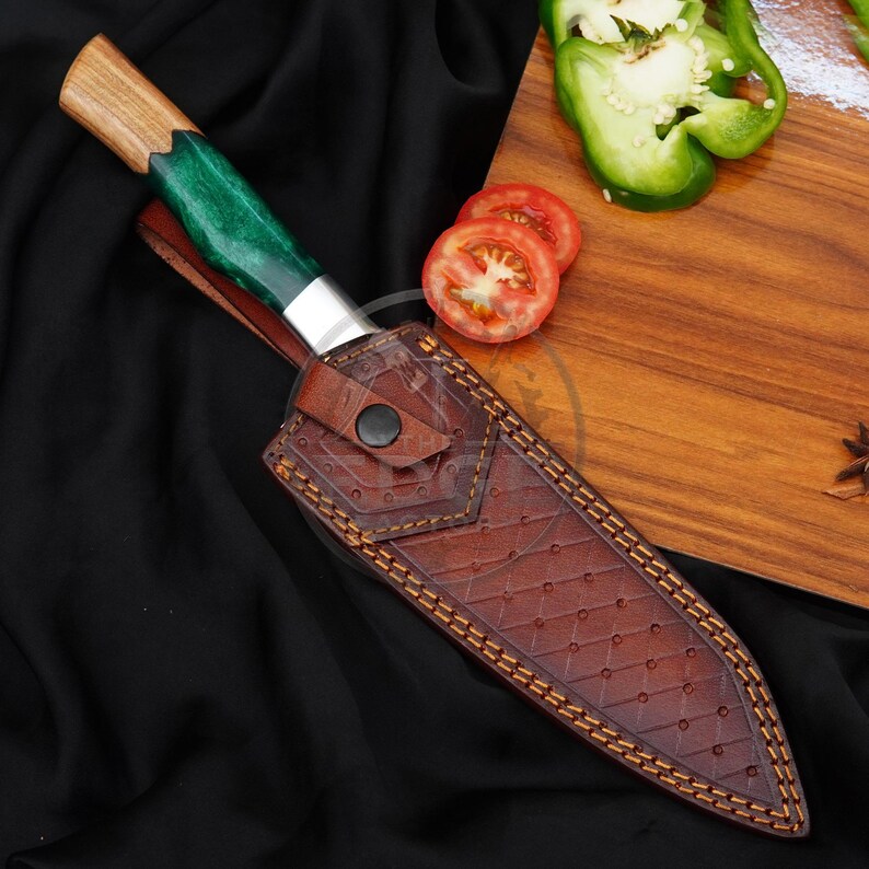 May include: A fixed-blade knife with a wooden and green resin handle, encased in a brown leather sheath featuring a diamond-patterned texture, a snap button closure, and visible stitching, positioned alongside sliced green bell pepper and tomato.