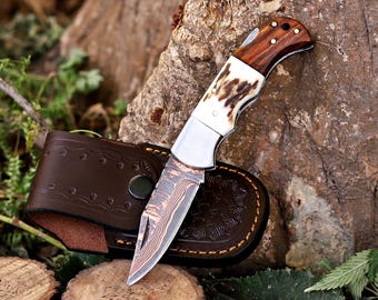 May include: A folding pocket knife with a Damascus steel blade, a white bone handle section, and a dark brown wood handle section with brass pins. The knife is positioned next to a dark brown leather sheath with decorative stitching and a snap closure.