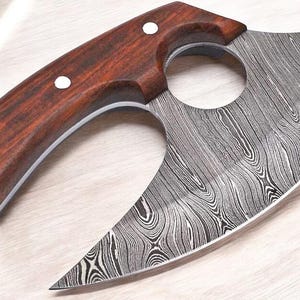 May include: A Damascus steel knife with a curved blade and a dark wood handle. The handle has three white dot accents. The blade has a unique wood grain pattern. The knife has a circular opening near the handle.