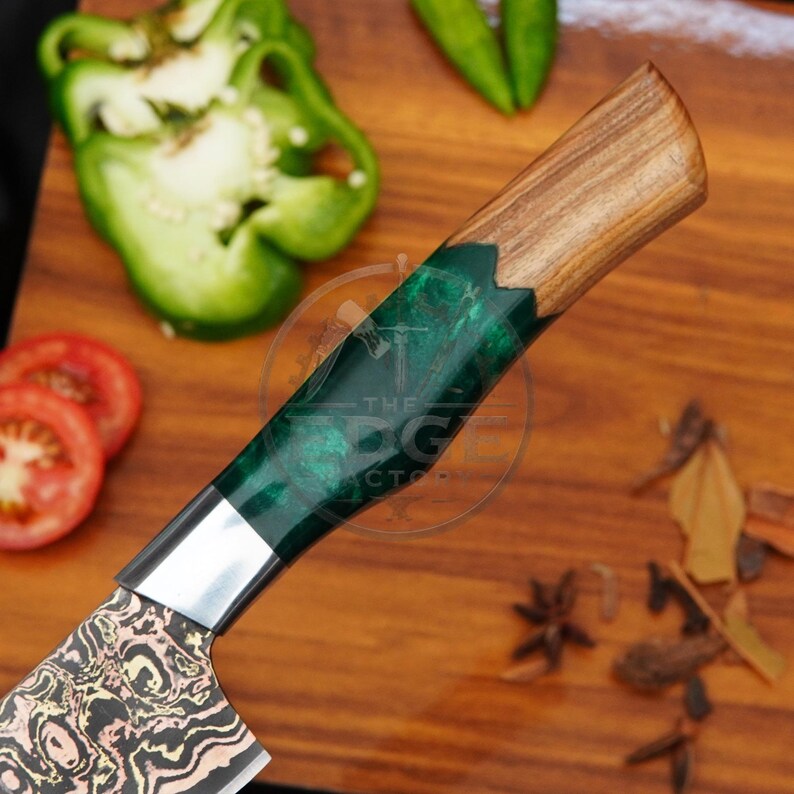 May include: A kitchen knife featuring a patterned metal blade and a handle constructed from a combination of natural wood and translucent emerald green resin. The handle displays a circular logo with the text THE EDGE FACTORY.