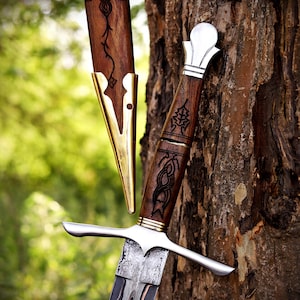 May include: A dagger with a dark wood handle featuring intricate black carvings, a polished silver crossguard, and a patterned metal blade. The matching wooden scabbard has a polished brass tip and a black engraved vine design on the wood.