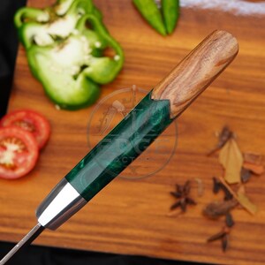 May include: A kitchen knife with a handle featuring a combination of natural wood grain at the top and a translucent emerald green resin section, separated by a silver-coloured metal bolster, with the text The Edge Factory visible on the handle.
