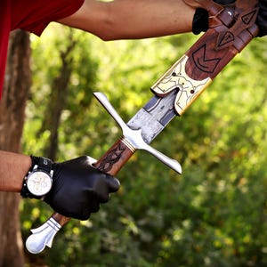 May include: A sword with a patterned steel blade, a wooden handle featuring carved details, and a silver-colored crossguard and pommel. The sword is partially inserted into a wooden scabbard adorned with decorative brass metal accents.