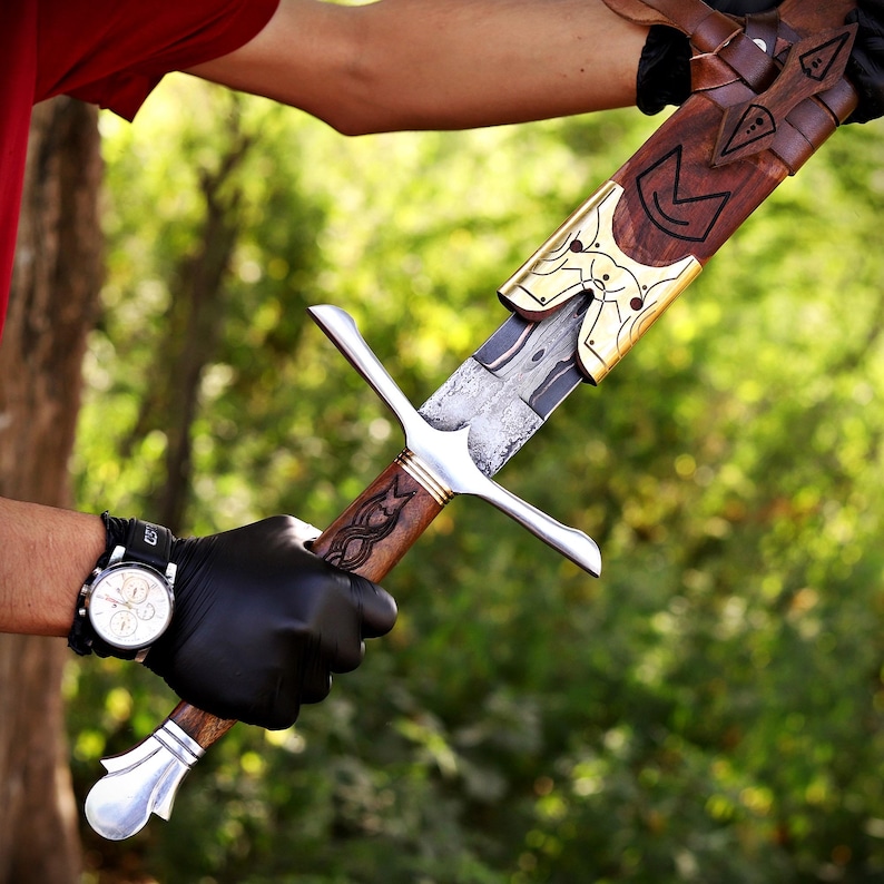 May include: A sword with a patterned steel blade, a wooden handle featuring carved details, and a silver-colored crossguard and pommel. The sword is partially inserted into a wooden scabbard adorned with decorative brass metal accents.