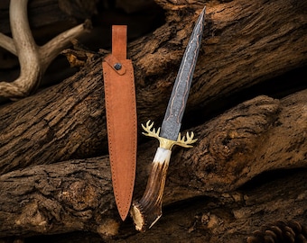 May include: A fixed-blade knife featuring a dark, patterned Damascus steel blade, a brass crossguard shaped like deer antlers, and a textured brown antler handle, accompanied by a tan leather sheath with a single metal rivet closure.