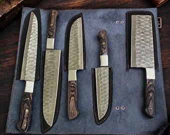 May include: A set of five kitchen knives with hammered metal blades and dark wood handles, each stored in a black leather sheath. The knives are arranged on a blue-grey leather roll-up mat featuring metal rivets and stitching along the edges.