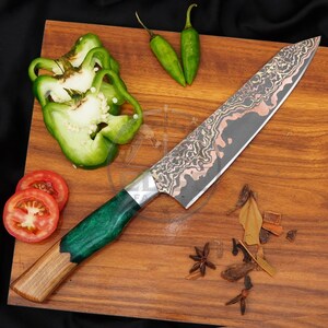 May include: A chef knife with a patterned Damascus steel blade featuring copper accents and a handle made of natural wood and green resin. The knife is displayed alongside sliced green bell peppers, two small green chilli peppers, and spices.