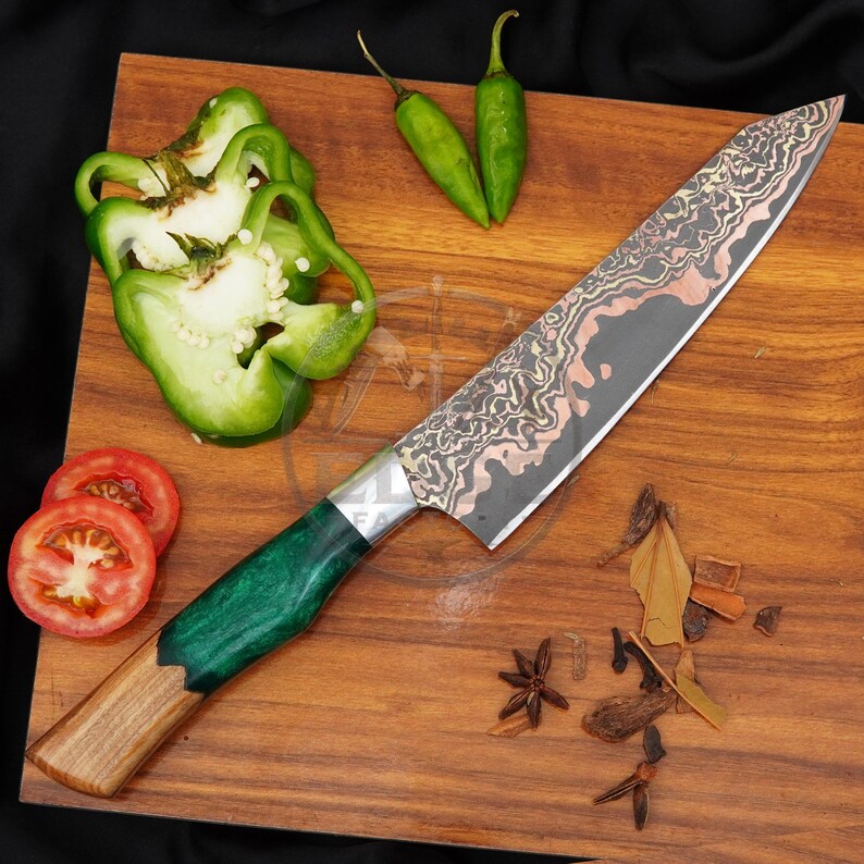 May include: A chef knife with a patterned Damascus steel blade featuring copper accents and a handle made of natural wood and green resin. The knife is displayed alongside sliced green bell peppers, two small green chilli peppers, and spices.